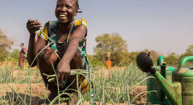 Eine Frau kniet lachend auf einem Gemüsebeet und hält eine Pflanze in der Hand.