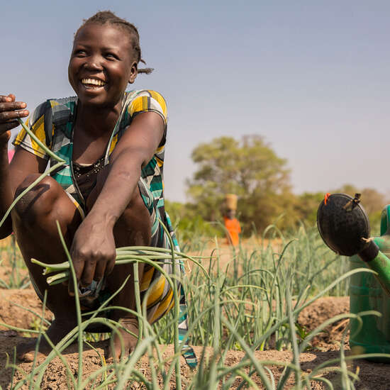 Eine Frau kniet lachend auf einem Gemüsebeet und hält eine Pflanze in der Hand.