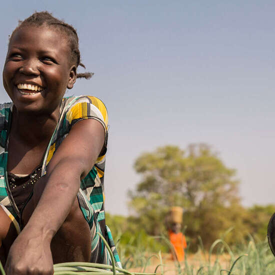 Eine Frau kniet lachend auf einem Gemüsebeet und hält eine Pflanze in der Hand.