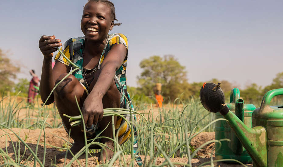 Ihre Hilfe für den Südsudan - jetzt spenden. Bild: Eine Frau kniet lachend auf einem Gemüsebeet und hält eine Pflanze in der Hand.