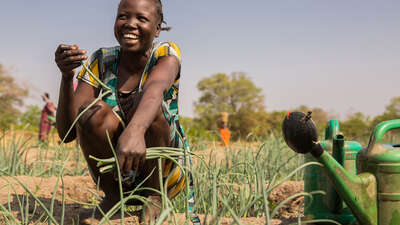 Eine Frau kniet lachend auf einem Gemüsebeet und hält eine Pflanze in der Hand.