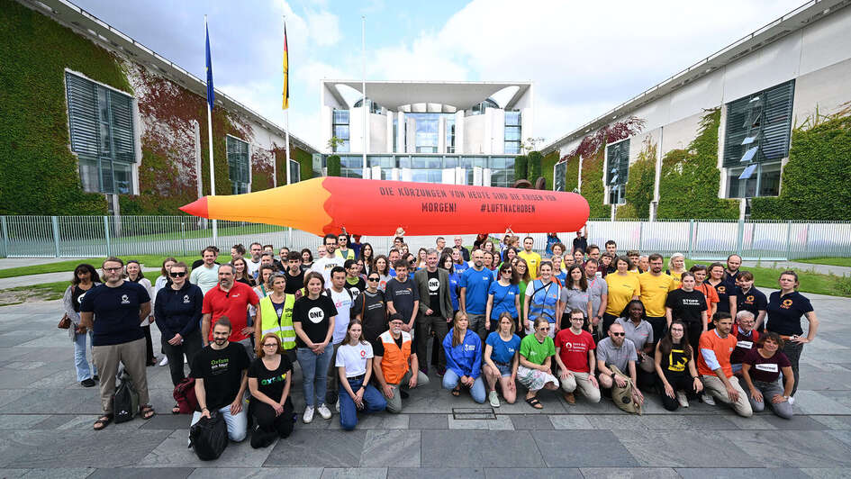 Gruppenfoto von demonstrierenden Personen von verschiedenen Hilfsorganisationen. Im Hintergrund das Kanzleramt und ein großer aufblasbarer Rotstift.