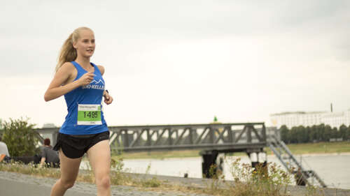 Eine junge Frau läuft am Rhein entlang. An ihrem blauen Oberteil ist eine Startnummer des Zero Hunger Run befestigt.