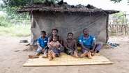 Ein Mann, eine Frau und drei Kinder sitzen auf einer Bastmatte, hinter ihnen eine Hütte aus Planen.