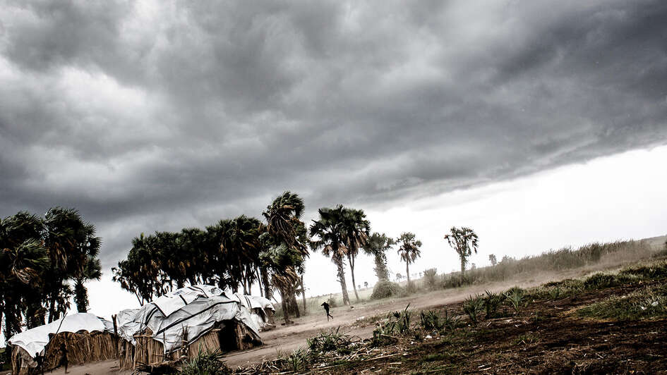 Dunkle Wolken am Himmel, neben aus Holz und Planen gebaute Häuser stehen Palmen, die sich im Wind beugen.