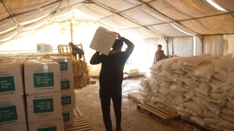 2019-nahrungsmittelhilfe-nord-aleppo-verteilung-syrien_C_welthungerhilfe.jpg Im Innern eines Zeltes: Links stehen aufgereihte und gestapelte Pakete der Welthungerhilfe. In der Mitte ein Mann von hinten, der ein Paket auf seinen Schultern trägt.