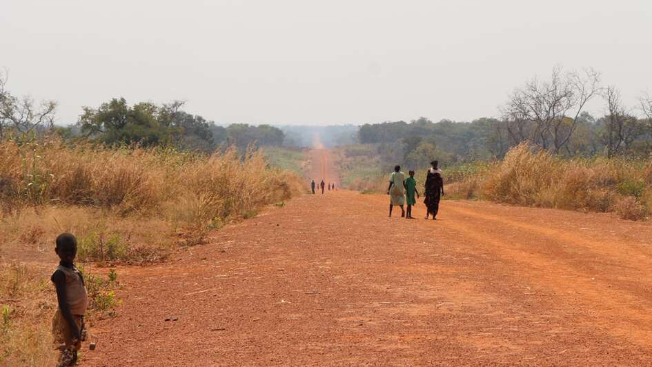 Eine unbefestigte Straße, die durch eine Savanne führt. Einige Menschen gehen auf ihr.
