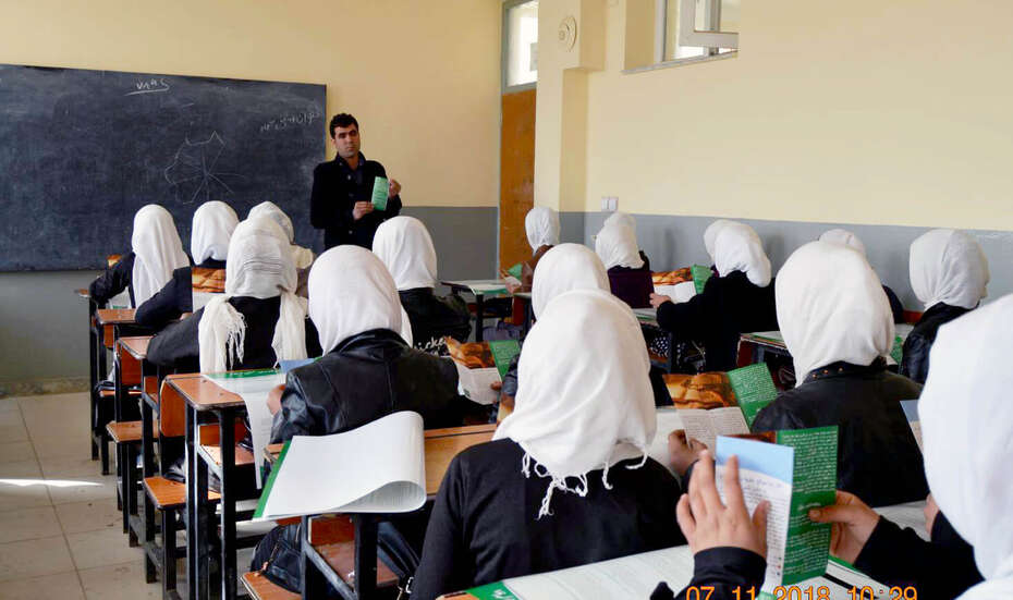Viele Frauen sitzen auf Schulbänken, vorne spricht ein Mann.