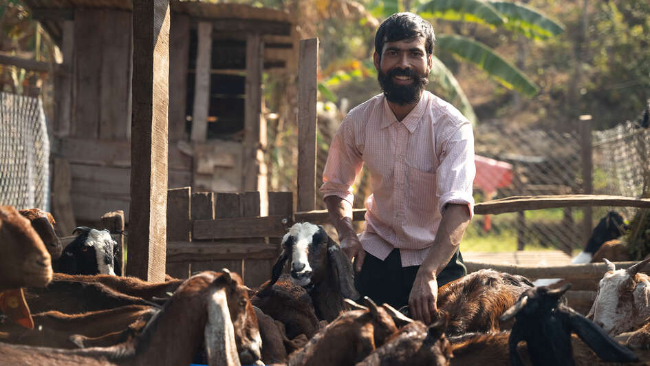 Mann lächelnd im Ziegenstall mit seinen Tieren im Hintergrund.