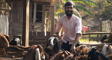 Mann lächelnd im Ziegenstall mit seinen Tieren im Hintergrund.
