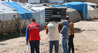 Vier Personen, zwei von ihnen in Uniformen von Hilfsorganisationen, in einem Flüchtlingslager in Syrien. Die Umgebung besteht aus provisorischen Unterkünften, die mit blauen und weißen Planen bedeckt sind.