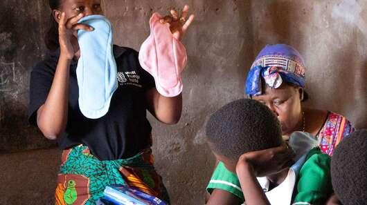 Eine Frau hält zwei Stoffbinden in der Hand, daneben sitzen mehrere Schülerinnen.