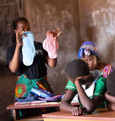 Eine Frau hält zwei Stoffbinden in der Hand, daneben sitzen mehrere Schülerinnen.
