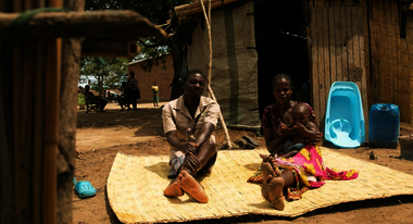 Auf einer Bastmatte sitzt ein Mann und eine Frau, die Frau hat ein Baby auf ihrem Schoß.