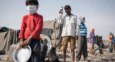 Mehrere Menschen, darunter auch Kinder, mit Mundschutz stehen in einer Schlange. Sie alle haben eine Metall-Schüssel in der Hand.