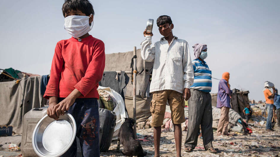 Mehrere Menschen, darunter auch Kinder, mit Mundschutz stehen in einer Schlange. Sie alle haben eine Metall-Schüssel in der Hand.