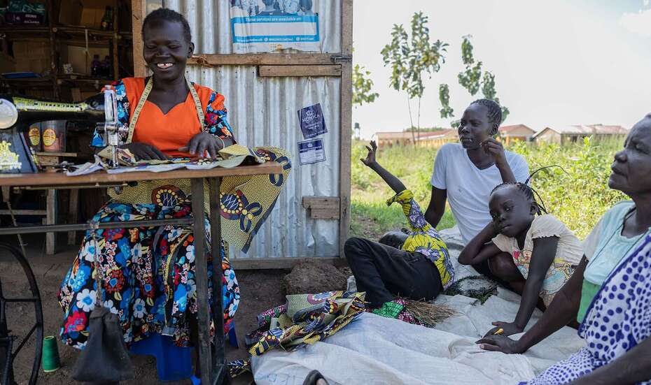 Frau arbeitet an Nähmaschine und lächelt, neben ihr sitzen mehrere Personen im Gespräch und schauen ihr zu
