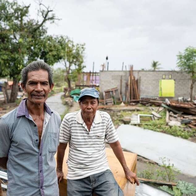 Zwei Männer schauen in die Kamera, hinter ihnen sieht man, was von den Häusern noch übrig geblieben ist.