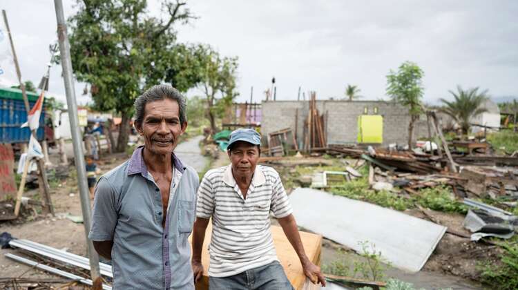 Zwei Männer schauen in die Kamera, hinter ihnen sieht man, was von den Häusern noch übrig geblieben ist.