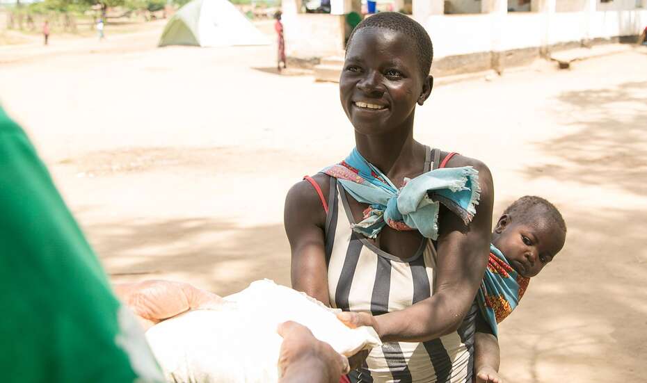 Eine Frau nimmt lächelnd ein kleines Päckchen entgegen. Auf dem Rücken trägt sie mit einem Tuch umschlugenes Baby.