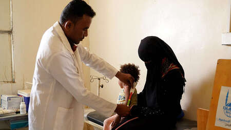 Ein Mann in weißen Kittel untersucht ein Kind, das auf dem Schoß seiner Mutter sitzt.