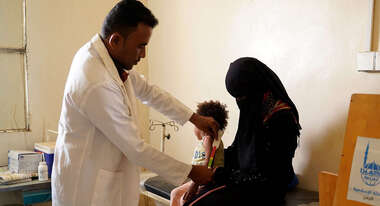 Ein Mann in weißen Kittel untersucht ein Kind, das auf dem Schoß seiner Mutter sitzt.