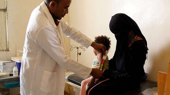 Ein Mann in weißen Kittel untersucht ein Kind, das auf dem Schoß seiner Mutter sitzt.