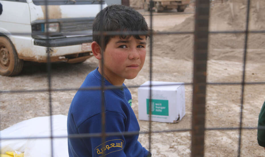 Ein Junge sitzt hinter einem Gitter und schaut in die Kamera. Im Hintergrund sieht man ein Welthungerhilfe-Paket.