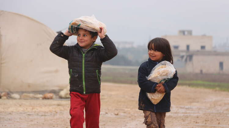 Ein Junge trägt eine Tüte Brot auf dem Kopf, ein Mädchen trägt eine Tüte Brot in den Armen. Im Hintergrund sind Gebäude und ein Zelt zu sehen.