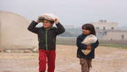 Ein Junge trägt eine Tüte Brot auf dem Kopf, ein Mädchen trägt eine Tüte Brot in den Armen. Im Hintergrund sind Gebäude und ein Zelt zu sehen.