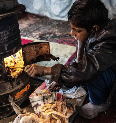 Ein Junge sitzt an einem Ofen und stochert mit einem Ast im Feuer.