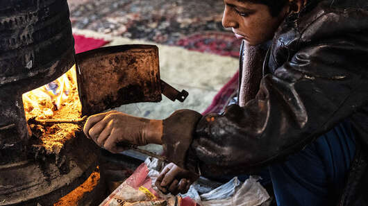 Ein Junge sitzt an einem Ofen und stochert mit einem Ast im Feuer.