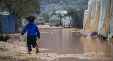 Ein kleines Mädchen von hinten, es läuft auf einem unbefestigten Weg, der überschwemmt ist. Im Hintergrund sieht man Zelte.