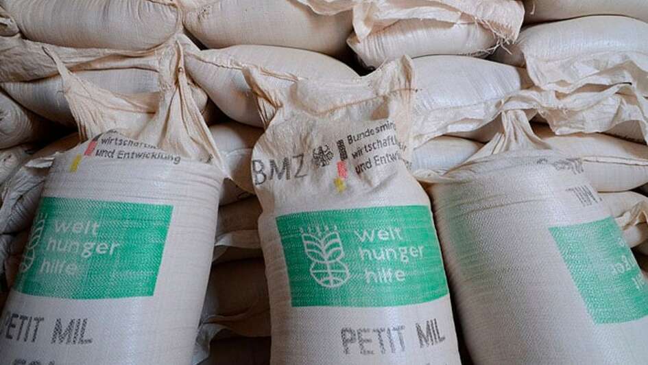 Drei vor Mehlsäcke der Welthungerhilfe stehen vor einem Sackstapel..