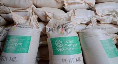 Drei vor Mehlsäcke der Welthungerhilfe stehen vor einem Sackstapel..