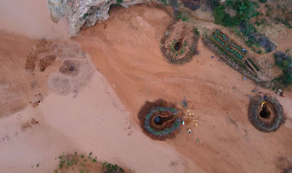 Luftaufnahme von einem ausgetrockneten Flussbett mit rötlicher Erde. Rechts sind drei Wasserlöcher zu sehen, um die herum etwas Grün wächst. 