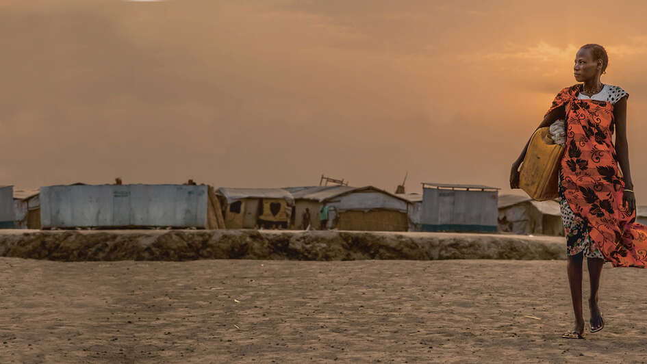 Im Vordergrund eine Frau mit einem gelben Kanister, im Hintergrund sieht man Zelte.