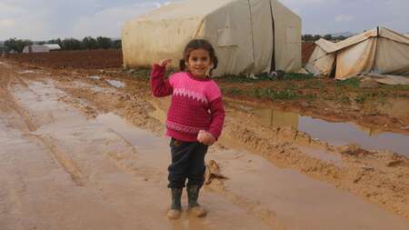 Ein lachendes Mädchen in Gummistiefeln steht auf einer matschigen Straße. Im Hintergrund sieht man ein weißes Zelt.