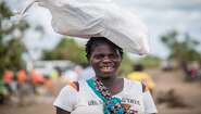 Eine Frau trägt ein Hilfsgüter-Paket auf dem Kopf und lächelt in die Kamera.