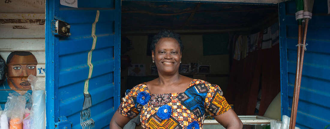 Eine Frau steht mit den Händen in den Hüften vor einem kleinen Laden, sie schaut lächelnd in die Kamera.