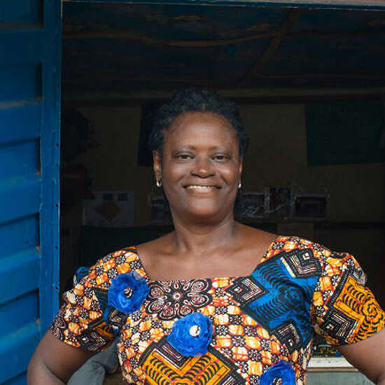 Eine Frau steht mit den Händen in den Hüften vor einem kleinen Laden, sie schaut lächelnd in die Kamera.