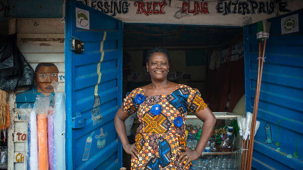 Eine Frau steht mit den Händen in den Hüften vor einem kleinen Laden, sie schaut lächelnd in die Kamera.