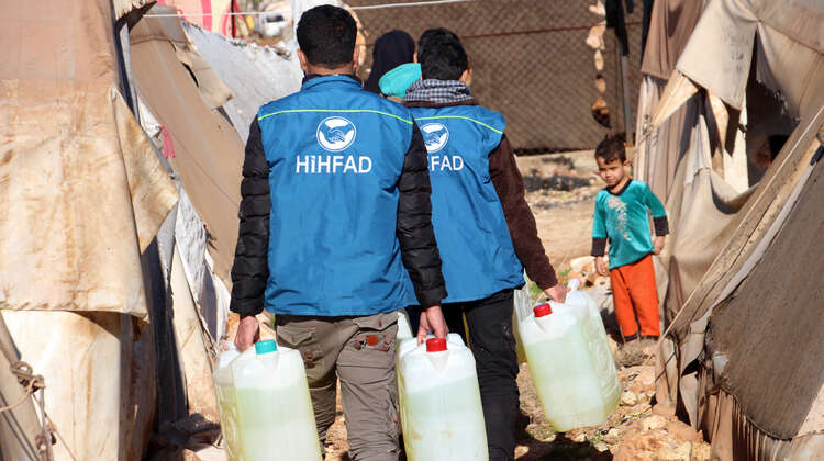 HIHFAD-Mitarbeiter in blauen Westen tragen Diesel in Kunststoffkanistern zu einem Zelt. Ein Kind schaut ihnen entgegen. 