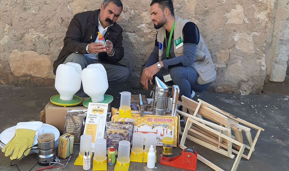 Zwei Männer sitzen an einer Wand. Vor ihnen sieht man mehrere Gegenstände.