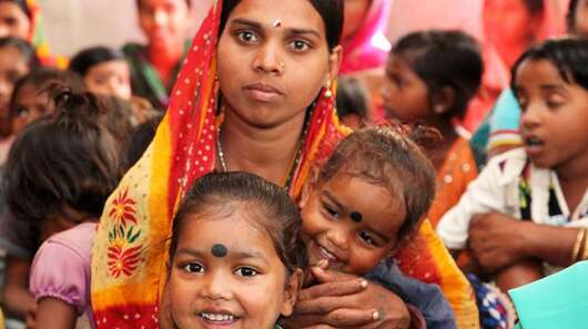 Eine Frau mit ihren zwei Kindern auf dem Schoss. Um sie herum sieht man viele andere Frauen.