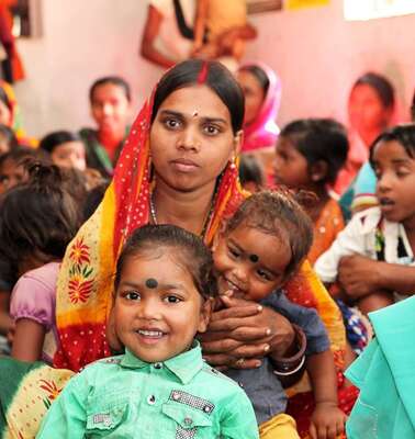 Eine Frau mit ihren zwei Kindern auf dem Schoss. Um sie herum sieht man viele andere Frauen.