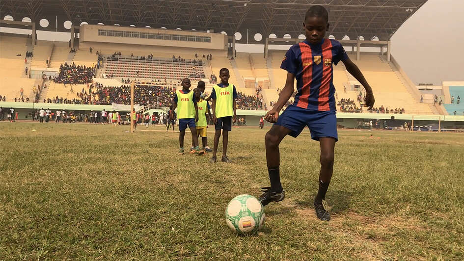 Ein Junge in Fußballtrikot und Ball im Vordergrund. Im Hintergrund weitere Kinder und die Tribüne des Stadions.