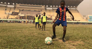 Ein Junge in Fußballtrikot und Ball im Vordergrund. Im Hintergrund weitere Kinder und die Tribüne des Stadions.