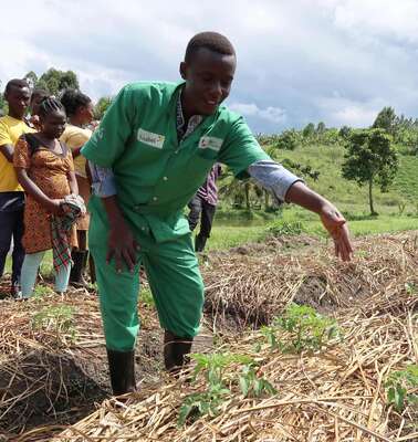 Eine Person in grünem Overall zeigt jungen Menschen etwas auf einem Gemüsebeet.
