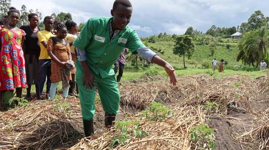 Ein Mann in grünem Overall zeigt jungen Menschen etwas auf einem Gemüsebeet.